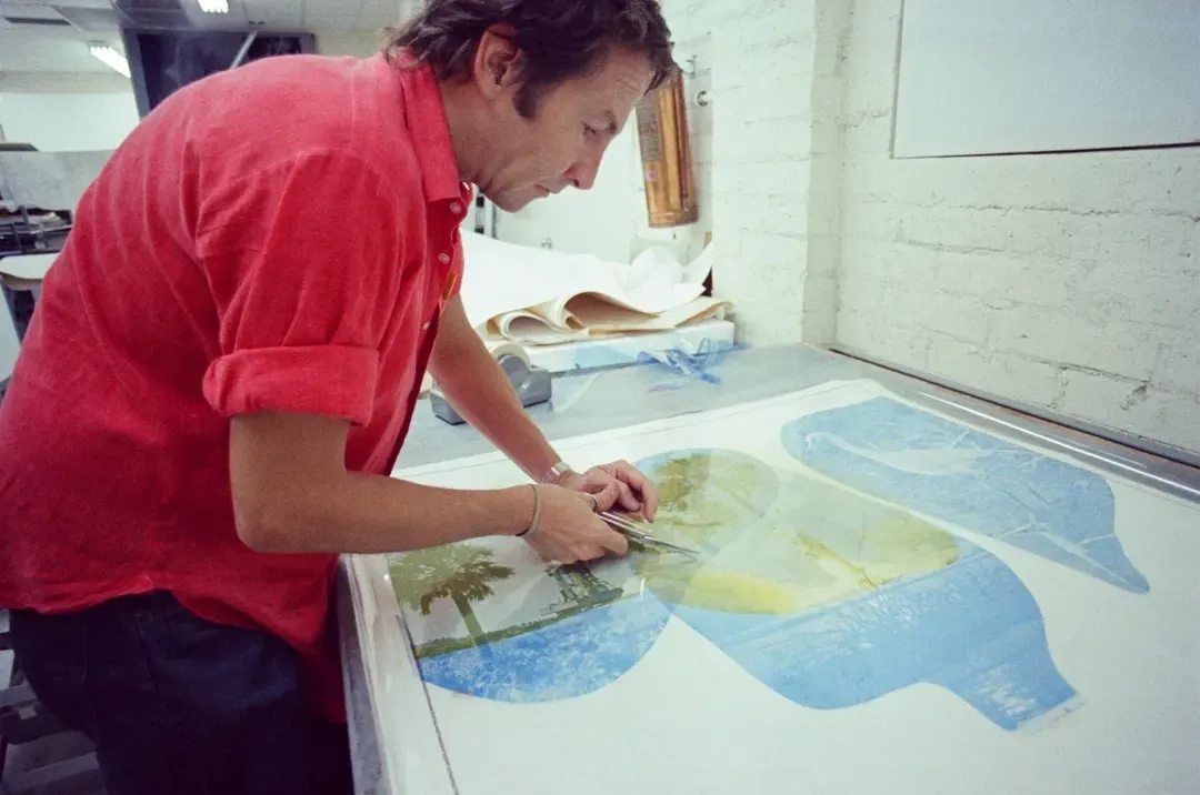 A man in a red shirt cutting into a large print on a lightbox in a print studio, working across overlapping blue and yellow shapes, one of them holding a photograph of palm trees.